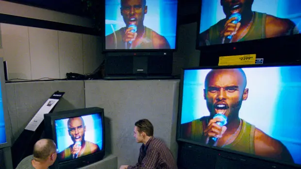 ca. 2001, Bloomington, Minnesota, USA --- Two men squat, looking at big screen televisions while shopping at Sears inside the Mall of America. A singer's face is shown on all of the TV screens. --- Image by © Layne Kennedy/Corbis