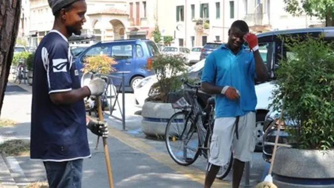 Curvi sotto il sole di una domenica d'agosto, a pulire le erbacce dalle strade, Friday e Celestian, nigeriani di 25 e 28 anni, cercano di mostrare l'altra faccia dell'immigrazione. Quella di chi vuole integrarsi nel Paese "onestamente", scrivono nel foglio di 'presentazione', appoggiato a terra. Da alcuni mesi lavorano gratis per la città di Padova - perchè non gliel'ha chiesto nessuno - e contano sulle offerte dei passanti. Nessuno dei due chiede l'elemosina. ANSA/GALVAN