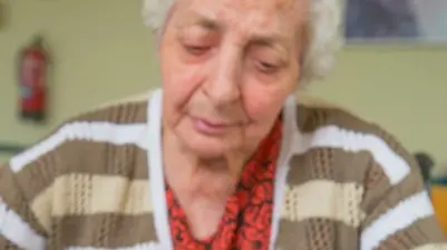 Old woman in a nursing home, on her one hundred birthday, blowing birthday's candles on a cake held by a man's hands.