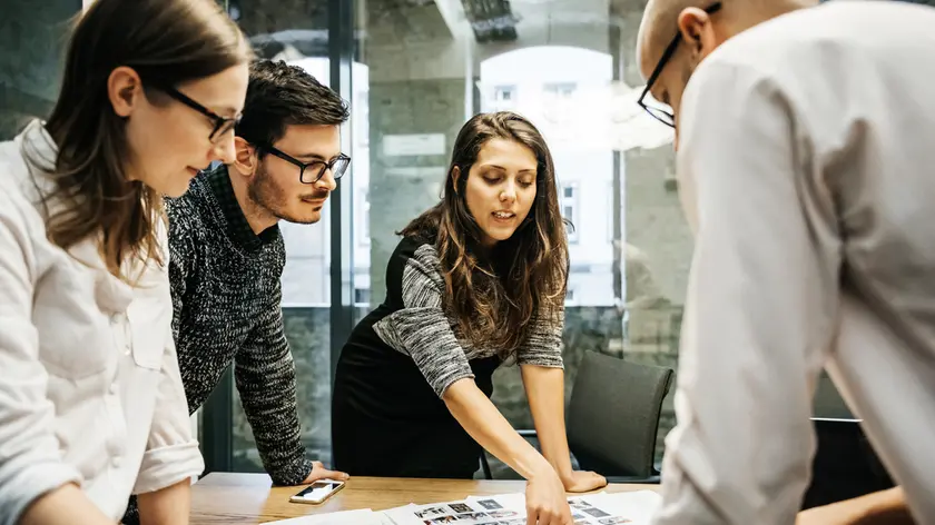 A team of young people in a modern office is discussing their project. The group consists of two men and two women, one of the is pointing at some papers while the others pay attention.