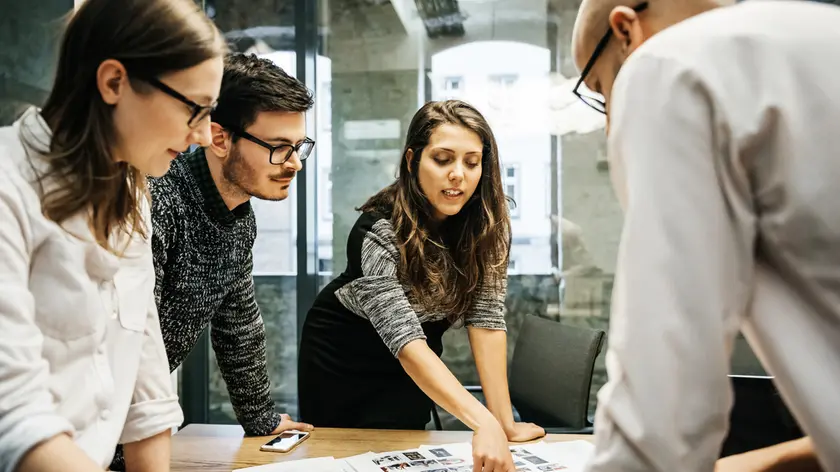 A team of young people in a modern office is discussing their project. The group consists of two men and two women, one of the is pointing at some papers while the others pay attention.